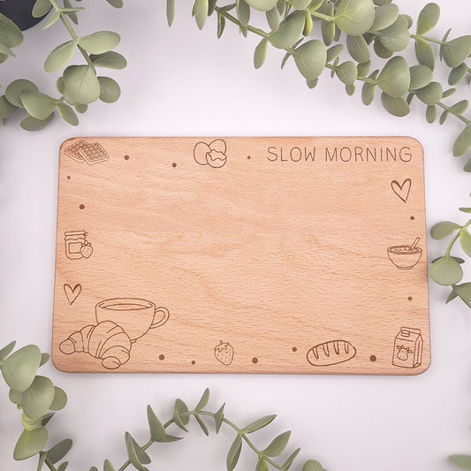 Draufsicht auf ein rechteckiges Holzbrettchen aus Buche mit abgerundeten Ecken. Die Gravur zeigt den Schriftzug 'Slow Morning' sowie verspielte Frühstücks-Icons wie Eier, Müsli, Milch und Gebäck in einem modernen Line-Art-Stil.