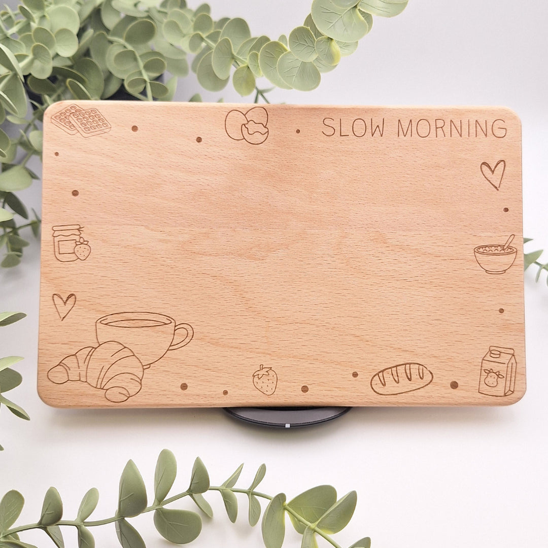 Hochwertiges Frühstücksbrettchen aus hellem Buchenholz mit fein detaillierter Lasergravur 'Slow Morning', umgeben von Eukalyptuszweigen. Sichtbar sind Motive wie Croissant, Kaffeetasse und Erdbeeren auf massiver Holzoberfläche.
