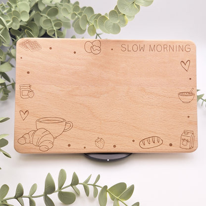 Hochwertiges Frühstücksbrettchen aus hellem Buchenholz mit fein detaillierter Lasergravur 'Slow Morning', umgeben von Eukalyptuszweigen. Sichtbar sind Motive wie Croissant, Kaffeetasse und Erdbeeren auf massiver Holzoberfläche.