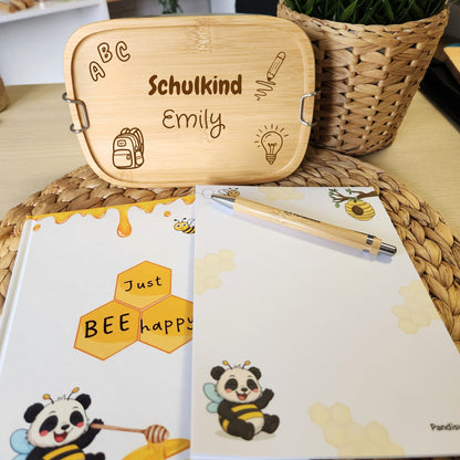 Einschulungsgeschenk-Set von Pandiswelt mit gravierter Brotdose, Notizbuch "Just Bee Happy" und passendem Notizblock, dekoriert auf einem Tisch