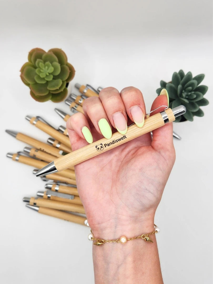 Ein hochwertiger Bambus-Kugelschreiber mit dem eingravierten Pandiswelt-Logo und Panda-Symbol, präsentiert in der Hand vor einem Stapel weiterer Holz-Kugelschreiber und grünen Sukkulenten