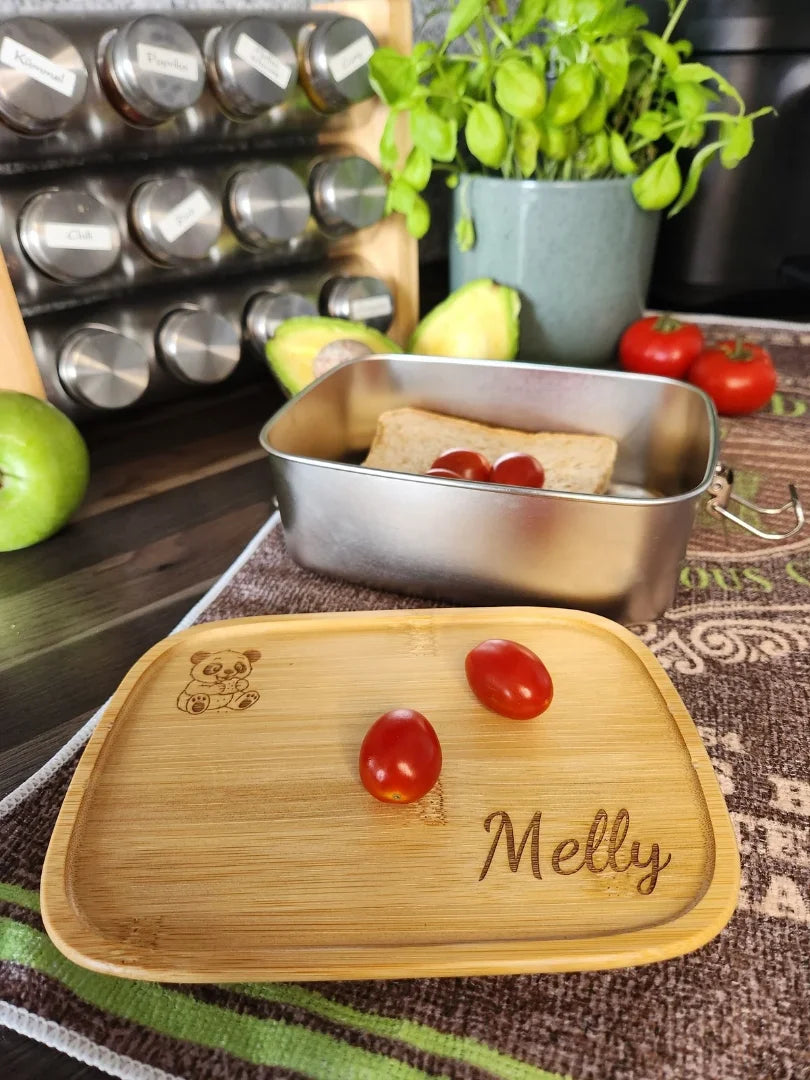 Funktionale Edelstahl-Brotdose, deren Bambusdeckel als praktisches Frühstücksbrettchen verwendet wird; zeigt die Lasergravur eines Pandas und aufgeschnittenes Gemüse in einer Küchenszenerie