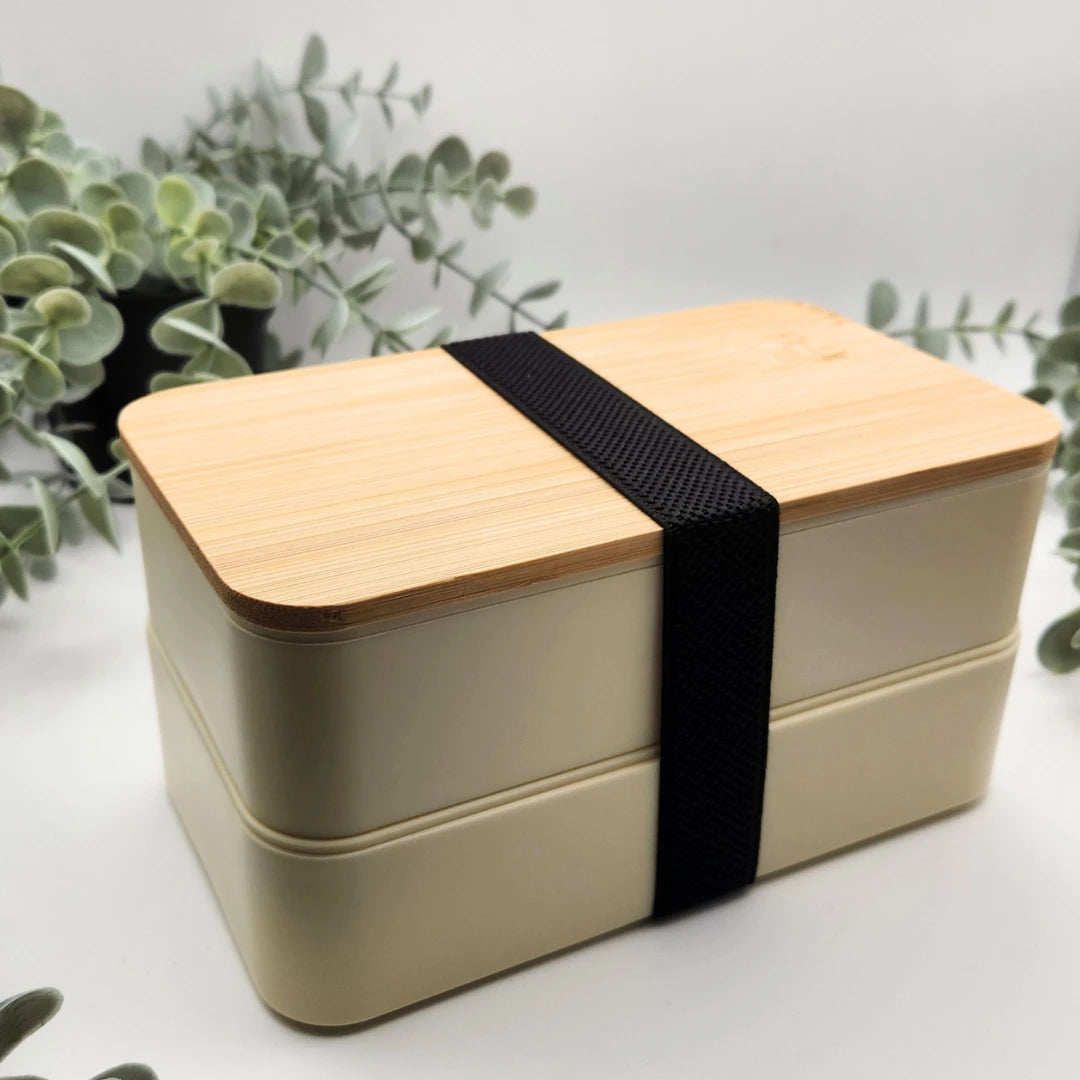 Geschlossene beige Brotdose mit zwei Etagen, Bambusdeckel und schwarzem Gummiband vor Eukalyptuszweigen