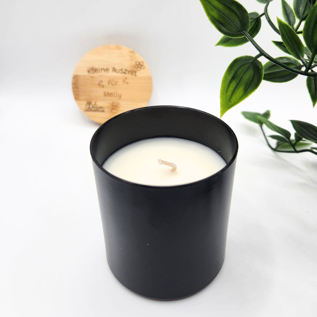Edle schwarze Duftkerze mit Vanille-Aroma in einem matten Aluminium-Tiegel; im Hintergrund ist der personalisierte Holzdeckel mit Gravur unter einem grünen Blatt-Zweig platziert
