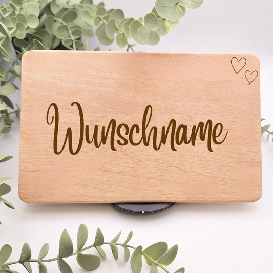 Frühstücksbrett aus Buchenholz mit der Gravur Wunschname in geschwungener Schreibschrift und zwei Herzen in der oberen Ecke, umgeben von Eukalyptuszweigen