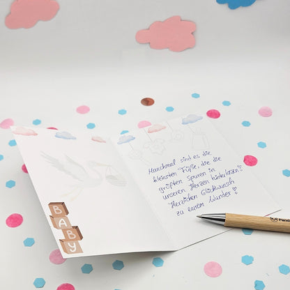 Innengestaltung der Geburtskarte mit Storch-Illustration und Baby-Bausteinen; zeigt ein handschriftliches Textbeispiel mit dem Spruch „Manchmal sind es die kleinsten Füße...“ und einen Bambus-Kugelschreiber