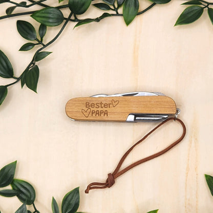 Ein personalisierbares Taschenmesser mit Bambus-Griff und der beispielhaften Gravur "Bester Papa" inklusive Herz-Symbolen und Lederschlaufe