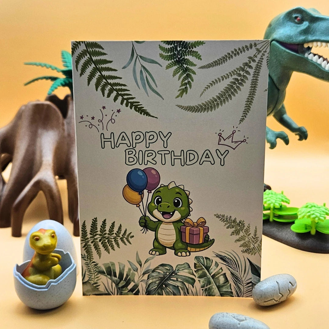 Detailansicht der Glückwunschkarte mit einem niedlichen grünen Dinosaurier, bunten Luftballons und Geschenk, umrahmt von Farnblättern im Jungle-Design