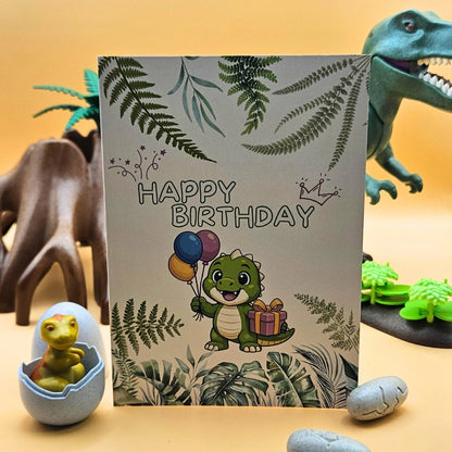 Detailansicht der Glückwunschkarte mit einem niedlichen grünen Dinosaurier, bunten Luftballons und Geschenk, umrahmt von Farnblättern im Jungle-Design