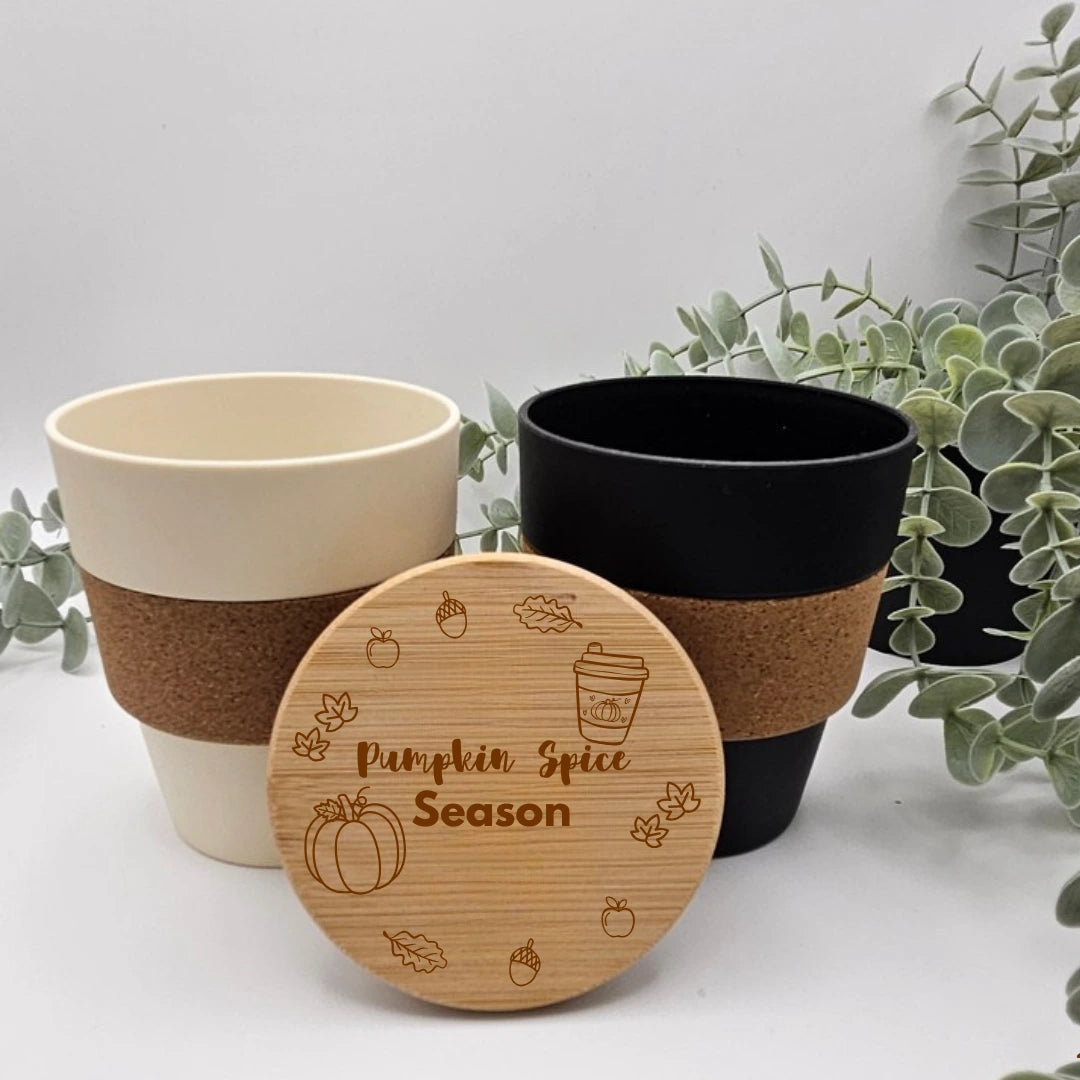 Kaffeebecher Herbstmotiv in Beige und Schwarz mit Korkmanschette, davor ein Bambusdeckel mit gravierter Herbstszene aus Kürbis, Laub und dem Schriftzug Pumpkin Spice Season