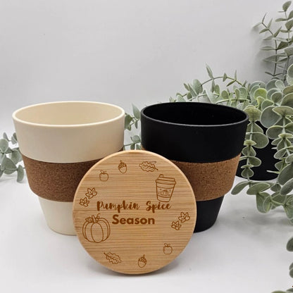 Kaffeebecher Herbstmotiv in Beige und Schwarz mit Korkmanschette, davor ein Bambusdeckel mit gravierter Herbstszene aus Kürbis, Laub und dem Schriftzug Pumpkin Spice Season