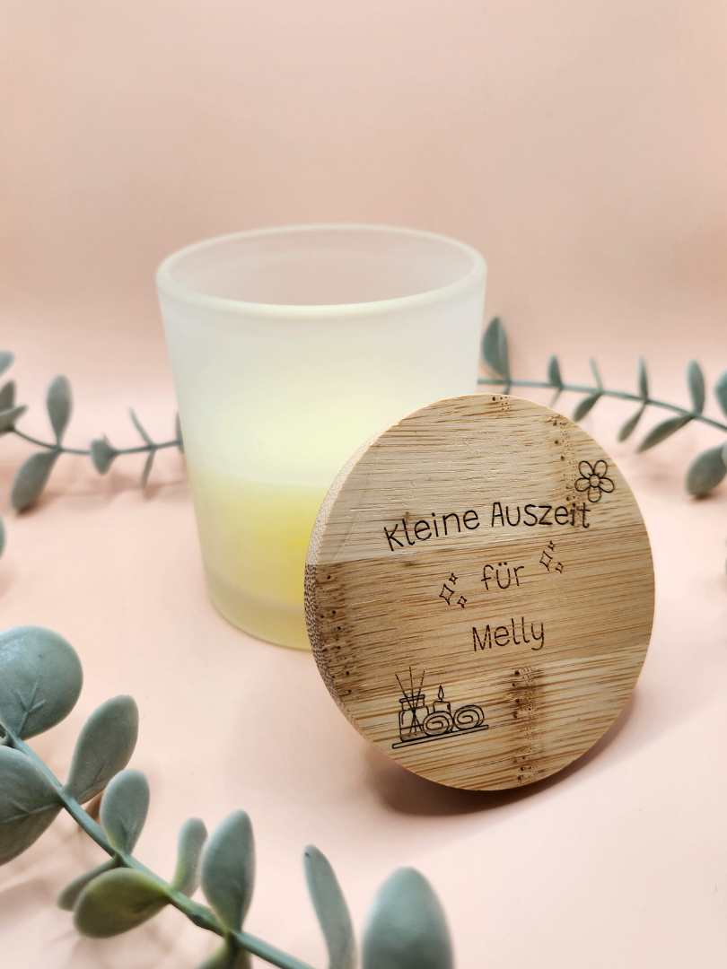 Nahaufnahme der personalisierten Kerze im satinierten Glas; der Holzdeckel lehnt am Glasrand und zeigt die individuelle Gravur „Kleine Auszeit für Melly“ mit feinen Spa-Illustrationen