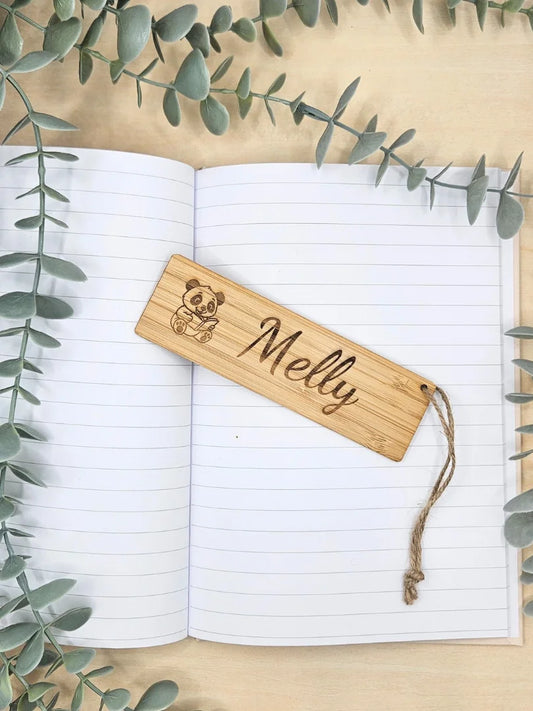 Ein personalisiertes Lesezeichen aus Bambus mit einer Panda-Gravur und dem beispielhaften Namen „Melly“, präsentiert auf einem aufgeschlagenen Notizbuch mit dekorativen grünen Zweigen
