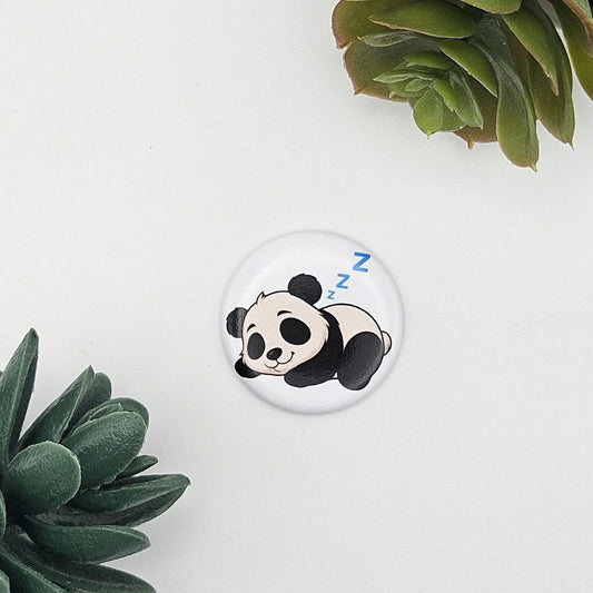 Detailansicht eines einzelnen runden Magnets (Ø 3,8 cm) mit der Illustration eines schlafenden Pandas und blauen „zzz“-Zeichen; dekorativ platziert auf weißem Grund zwischen grünen Sukkulenten-Zweigen