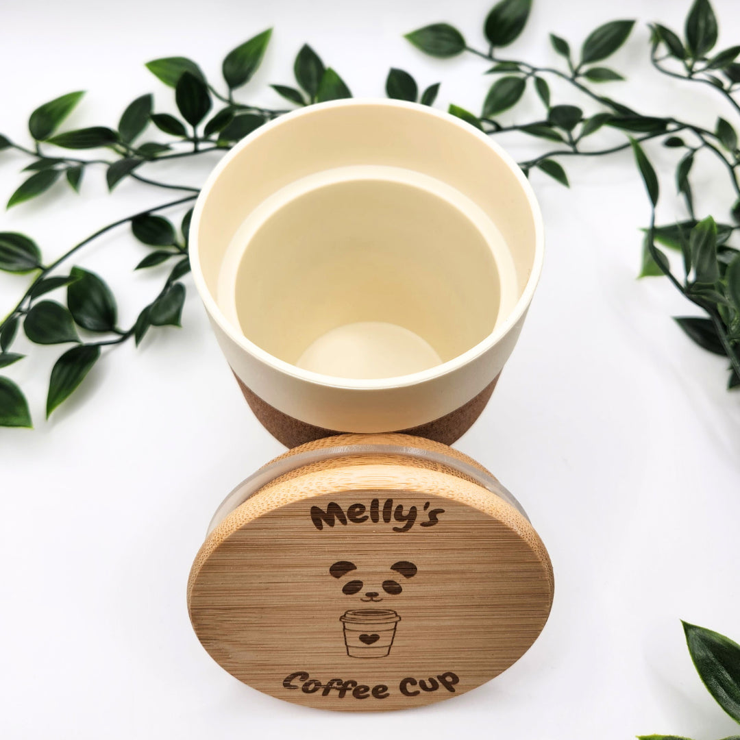 Nahaufnahme des geöffneten Mehrweg-Kaffeebechers mit abgenommenem Bambusdeckel; sichtbar ist die detailreiche Lasergravur „Melly's Coffee Cup“ vor einem natürlichen Blätter-Hintergrund