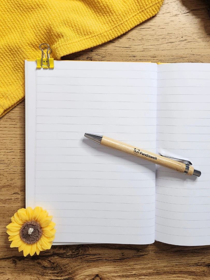 Geöffnetes liniertes A5 Notizbuch mit 64 Seiten und dem dazugehörigen Marken-Kugelschreiber aus Bambus; zeigt die hochwertige Papierqualität und das helle Design, ideal für tägliche Notizen und Journaling