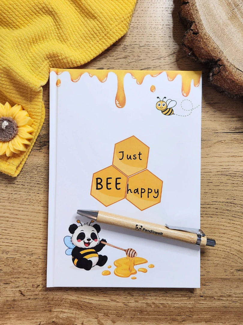 Notizbuch A5 Set von Pandiswelt mit dem Titel „Just BEE happy“ und einem Panda im Bienen-Kostüm; inklusive passendem Bambuskugelschreiber, dekoriert in einer sonnengelben Lifestyle-Umgebung mit Sonnenblumen-Kerze