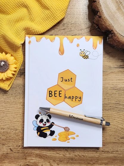 Notizbuch A5 Set von Pandiswelt mit dem Titel „Just BEE happy“ und einem Panda im Bienen-Kostüm; inklusive passendem Bambuskugelschreiber, dekoriert in einer sonnengelben Lifestyle-Umgebung mit Sonnenblumen-Kerze