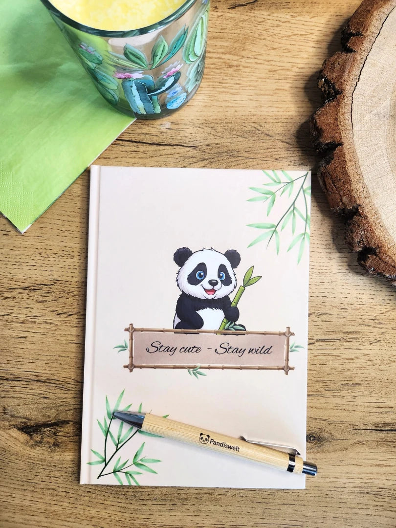 A5 Notizbuch-Set von Pandiswelt mit dem Titel „Stay cute - Stay wild“ und Panda-Motiv; inklusive hochwertigem Bambuskugelschreiber, ästhetisch präsentiert in einer grünen Lifestyle-Umgebung auf einem Holztisch