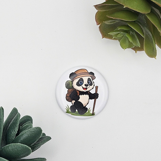 Einzelner runder Kühlschrankmagnet (Ø 3,8 cm) mit der Illustration eines wandernden Pandas mit Hut, Rucksack und Wanderstock; moderne Präsentation auf weißem Grund zwischen grünen Blättern
