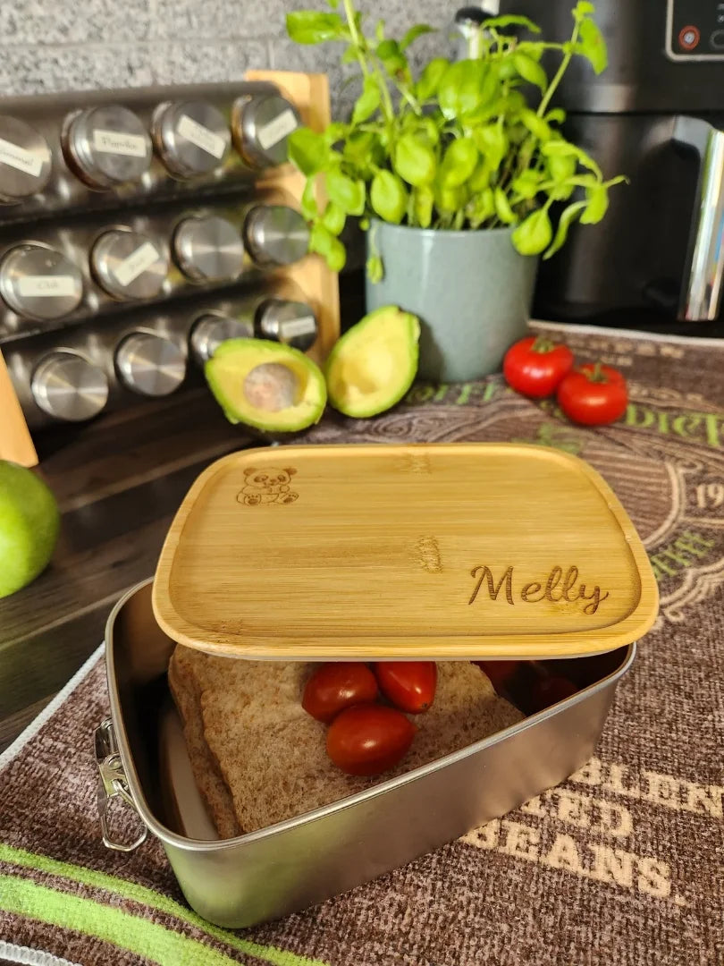 Personalisierte Pausenbox aus Edelstahl, gefüllt mit Brot und Tomaten; der gravierte Bambusdeckel zeigt beispielhaft den Namen „Melly“ und eignet sich perfekt für gesundes Vesper in der Schule
