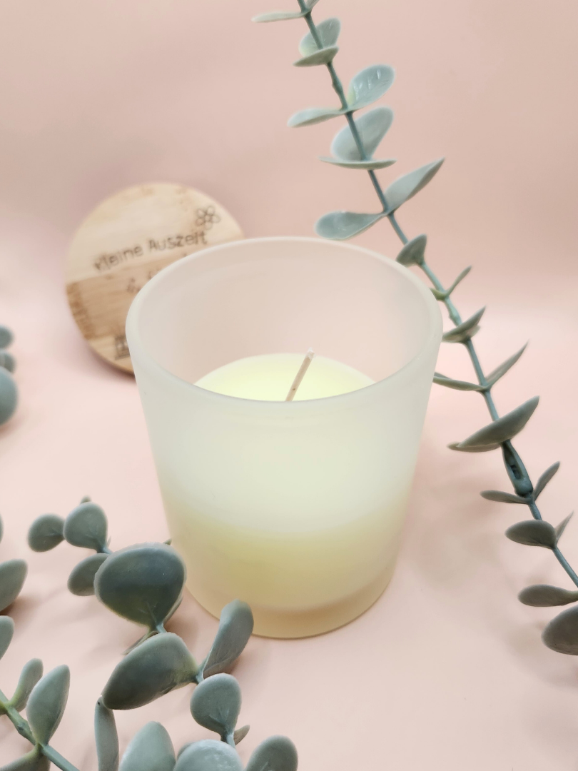Personalisierte Vanille-Duftkerze in einem edlen matten Milchglas vor zartrosa Hintergrund; der gravierte Bambusdeckel ist im Anschnitt sichtbar, dekorativ umrahmt von Eukalyptus-Zweigen