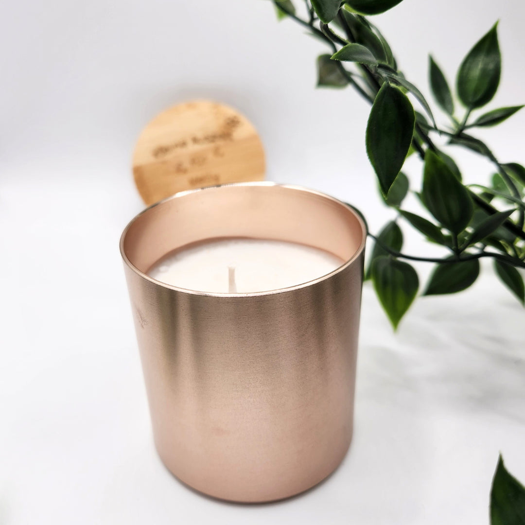Ästhetische Vanille-Duftkerze im rosegoldenen Metalltiegel mit weißem Wachs und Docht; im Hintergrund ist der gravierte Holzdeckel zwischen grünen Blättern für ein entspanntes Wellness-Ambiente angedeutet