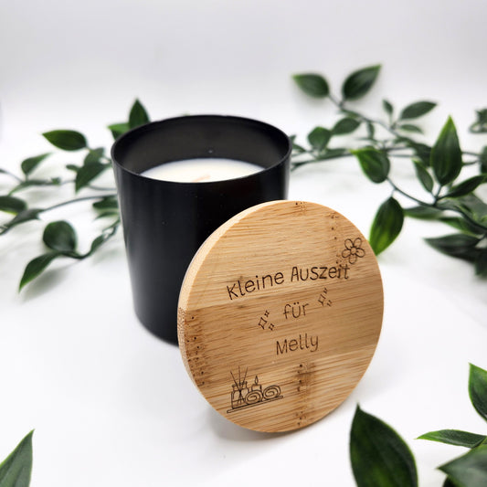 Nahaufnahme der schwarzen Vanillekerze; der gravierte Deckel lehnt am Tiegel und zeigt die individuelle Widmung „Kleine Auszeit für Melly“ sowie filigrane Spa-Motive