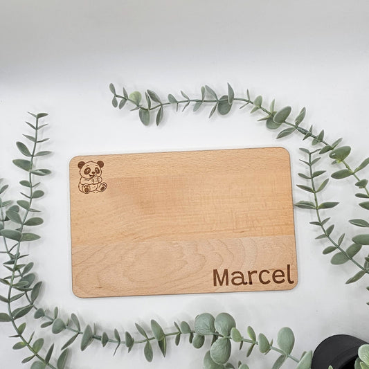Individuelles Frühstücksbrettchen aus massivem Buchenholz mit Lasergravur eines naschenden Pandas und dem beispielhaften Namen „Marcel“; hochwertiges Naturholz-Vesperbrett für Kinder, dekorativ umrahmt von grünen Blättern