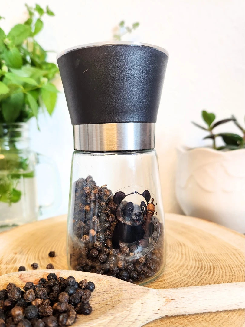 Befüllte Pfeffermühle aus Glas mit 100g ganzen schwarzen Pfefferkörnern auf einem rustikalen Holzbrett; das Gewürz-Set von Pandiswelt zeigt eine hochwertige Panda-Illustration und wird mit frischen Kräutern für die moderne Küche präsentiert