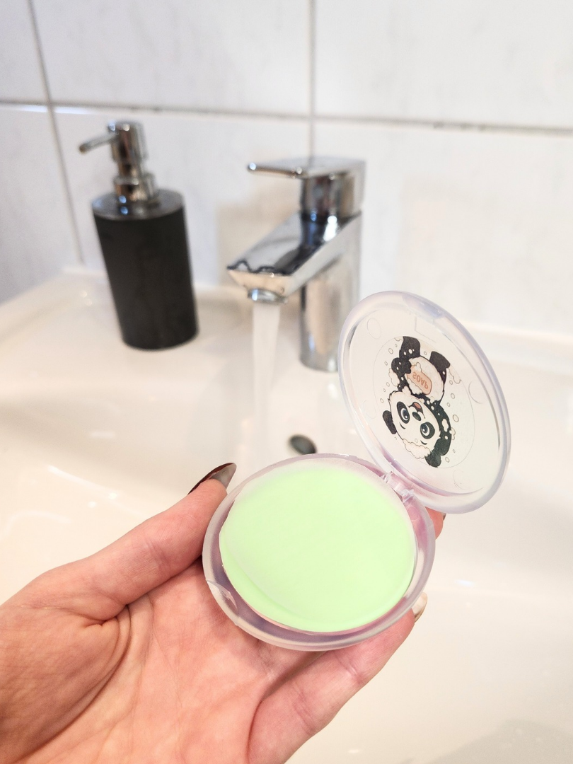 Geöffnete Panda-Seifendose mit grünen Seifenblättern in der Hand vor einem fließenden Wasserhahn; kompakte Lösung für saubere Hände beim Camping oder auf Ausflügen