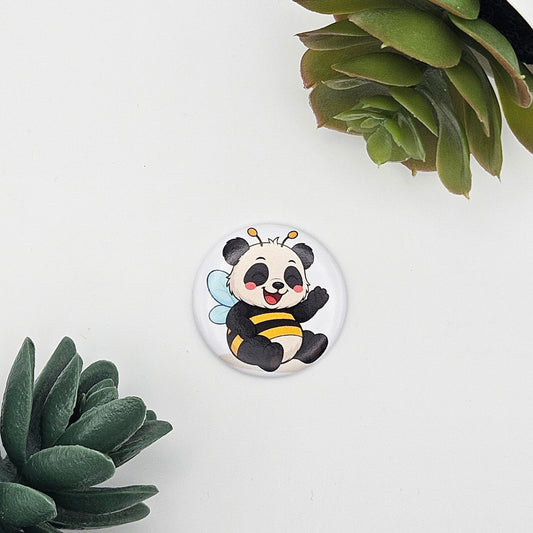 Einzelner runder Kühlschrankmagnet (Ø 3,8 cm) mit einer niedlichen Illustration eines Pandas im Bienen-Look; moderne Präsentation auf weißem Hintergrund zwischen grünen Sukkulenten-Blättern