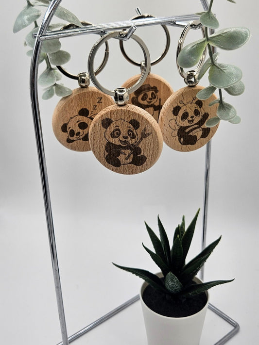 Verschiedene Panda-Schlüsselanhänger aus Holz an einem silbernen Halter; sichtbar sind Motive wie der schlafende Panda, der Wander-Panda und der Bienen-Panda, dekoriert mit Eukalyptus-Zweigen