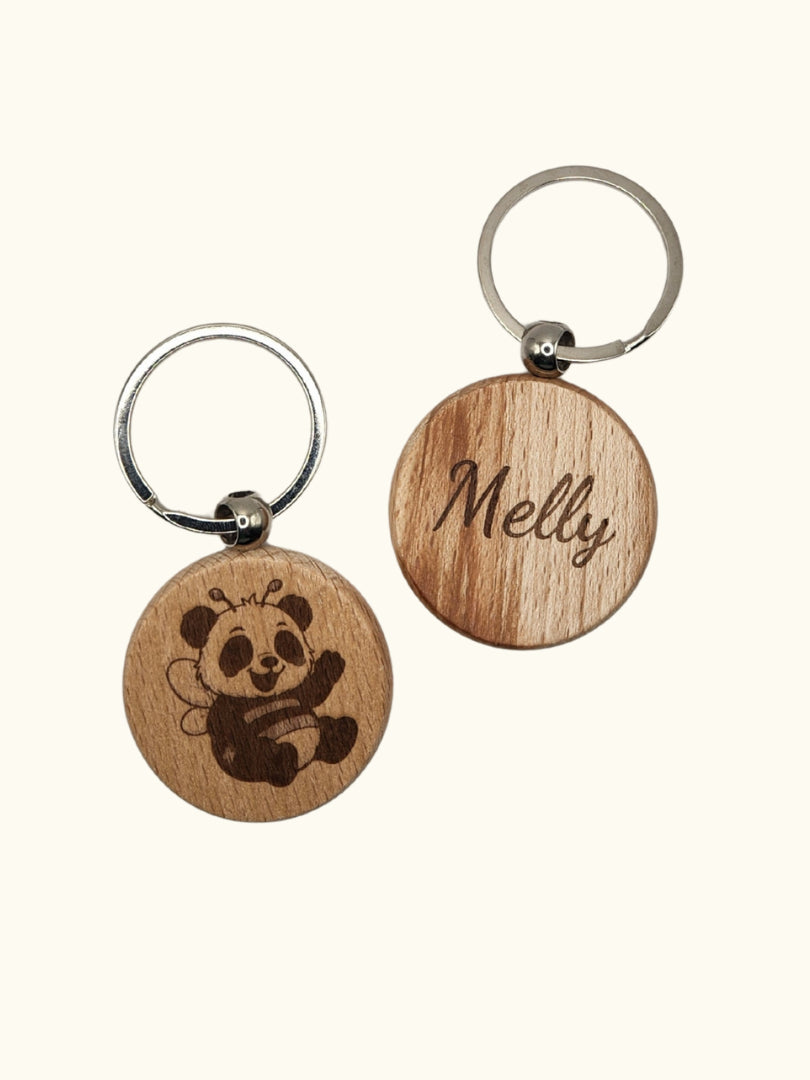 Personalisierter Schlüsselanhänger aus Buchenholz (Ø 4 cm) in der Ansicht von Vorder- und Rückseite; zeigt die Lasergravur eines Bienen-Pandas und die beispielhafte rückseitige Namensgravur „Melly“ auf hellem Hintergrund