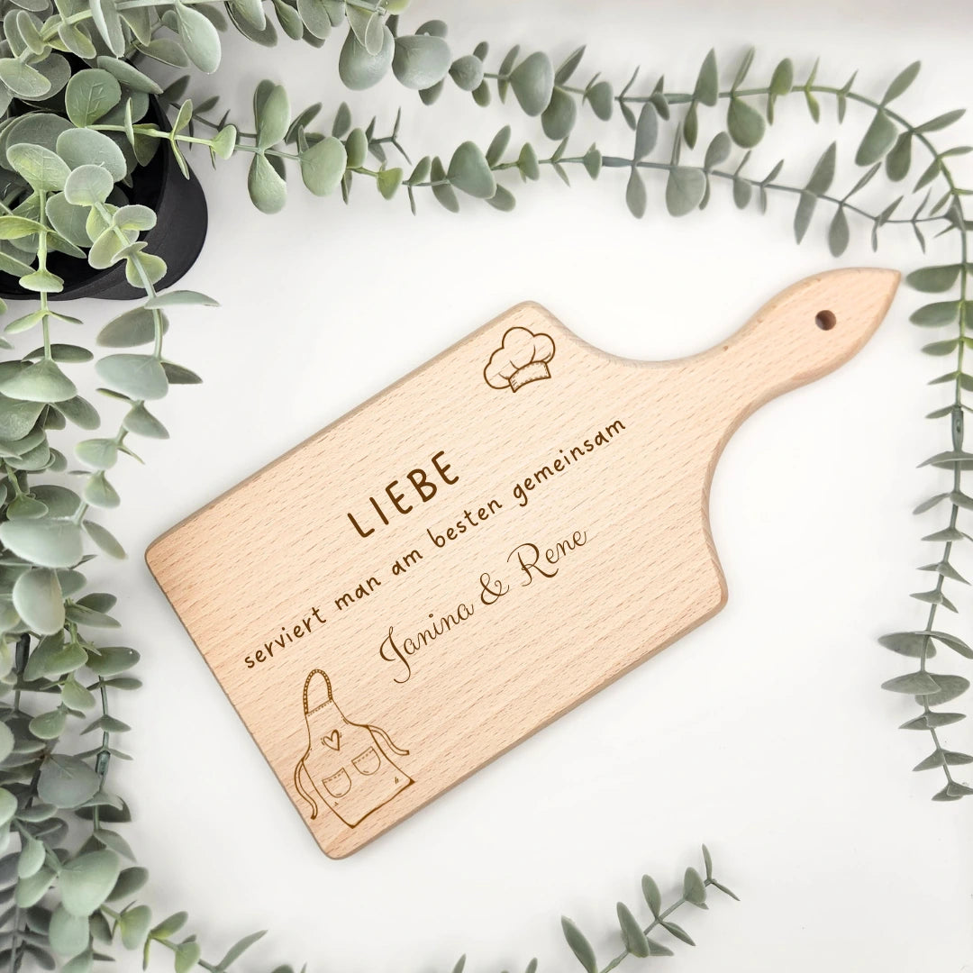 Personalisiertes Servierbrett als Einweihungsgeschenk mit Namen beispielsweise "Janina & Rene" und dem Text "Liebe serviert man am besten gemeinsam". Dazu eine Kochmütze und ein weiteres Motiv passend zur Küche.