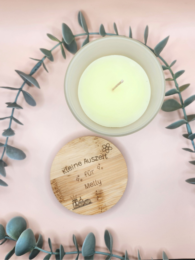 Ästhetische Draufsicht auf die geöffnete Vanillekerze im Glas und den danebenliegenden Deckel mit der Gravur „Kleine Auszeit für Melly“; platziert in einem Kranz aus frischem Eukalyptus